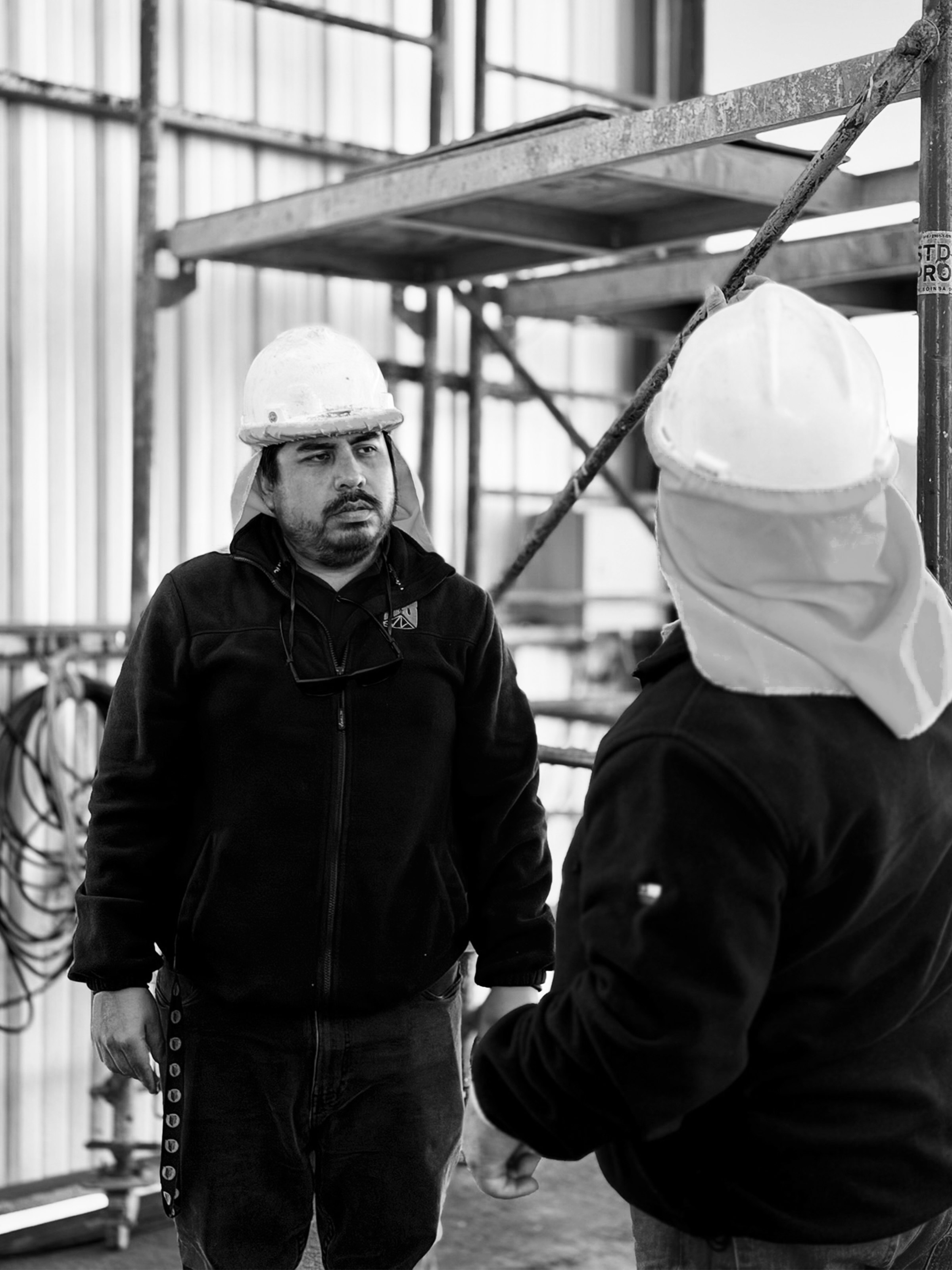 Dos trabajadores con cascos conversando en obra, estructura metálica de fondo